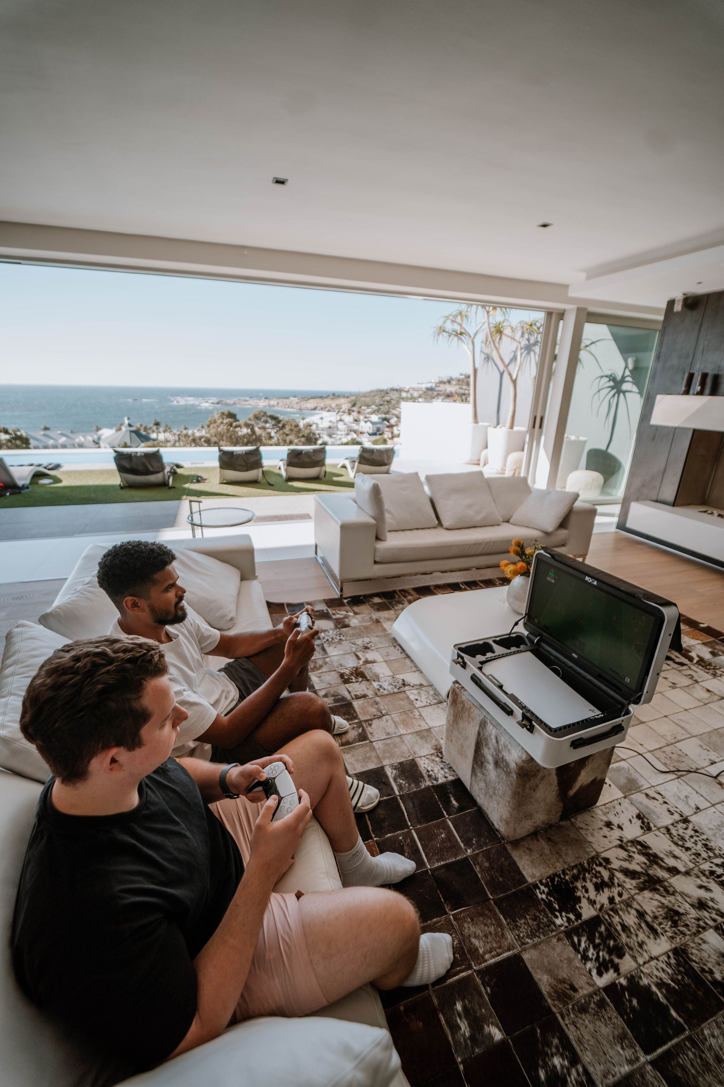 Two men sitting on a couch in a modern living room with a view of the ocean playing on a portable gaming  case